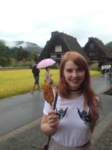Ingvill, jente med langt hår, som holder en frityrstekt noe på pinne foran japanske hus